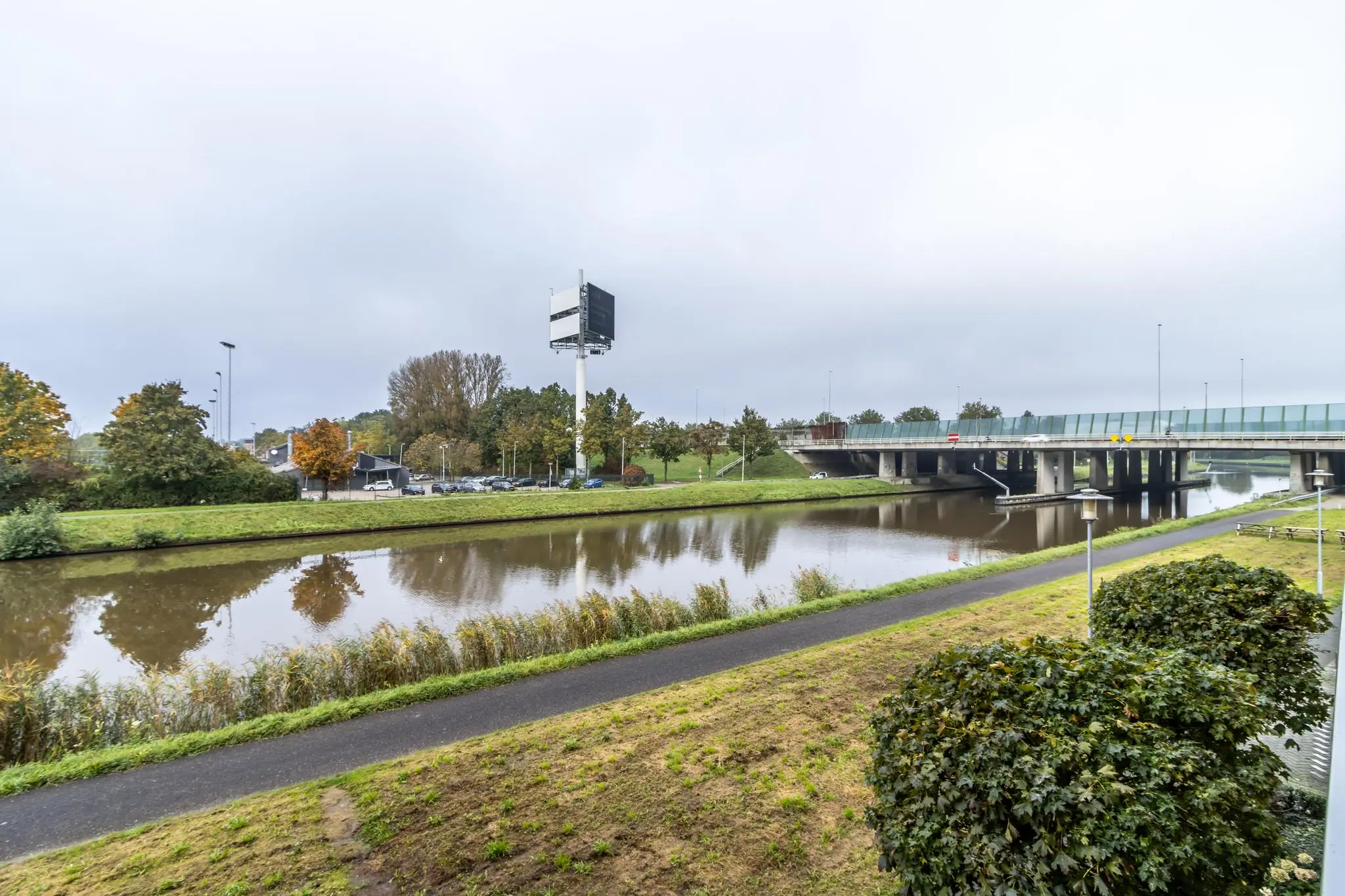 Brug over het kanaal langs de Lage Biezenweg met op de achtergrond een groot reclamebord en herfstige bomen.